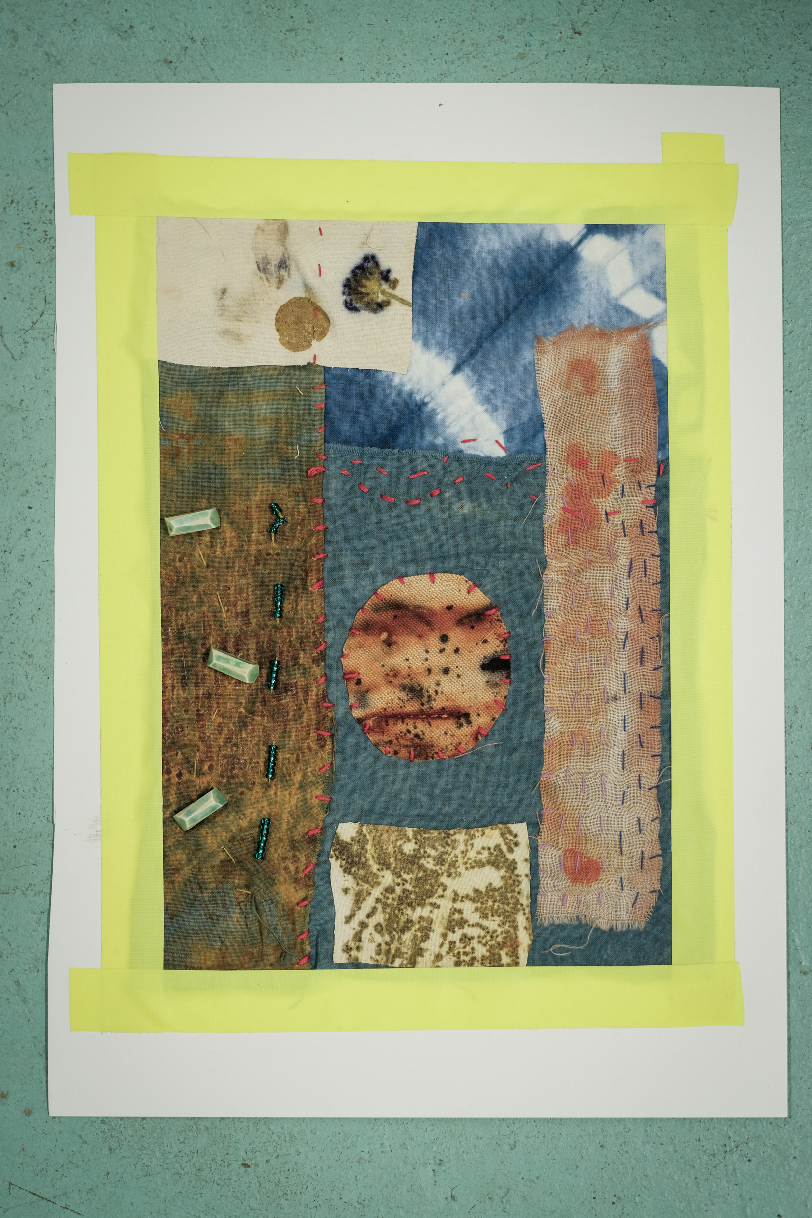 Patchwork textile art with various fabric swatches in blue, brown, beige, and orange, embroidered with red and blue stitches and adorned with green beads, mounted on white paper framed with yellow tape.